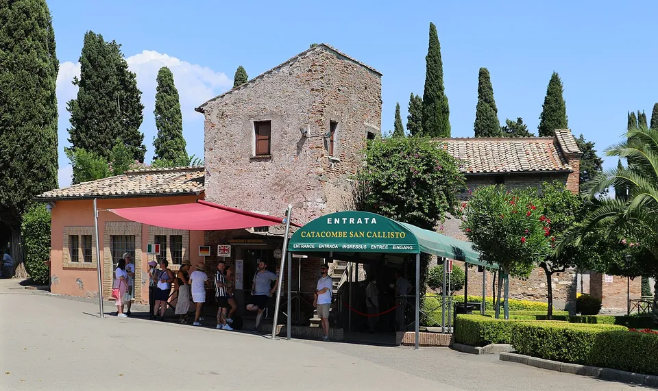 A low pink building and an older stone house among cypresses, with a green awning reading "Entrata Catacombe San Callisto" over a doorway where visitors are queuing.