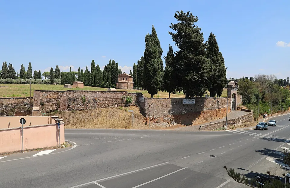 A road junction in bright sun, beyond it a long wall of old brick and stone with a gate in it, and behind the wall a field with tall cypresses and a few small brick buildings.