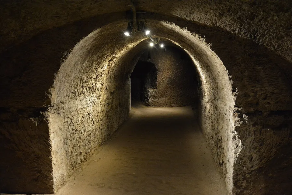 A long, low barrel-vaulted corridor cut into rough rock, lit by a row of small lamps along the ceiling, with a level earth floor running away into the dark.