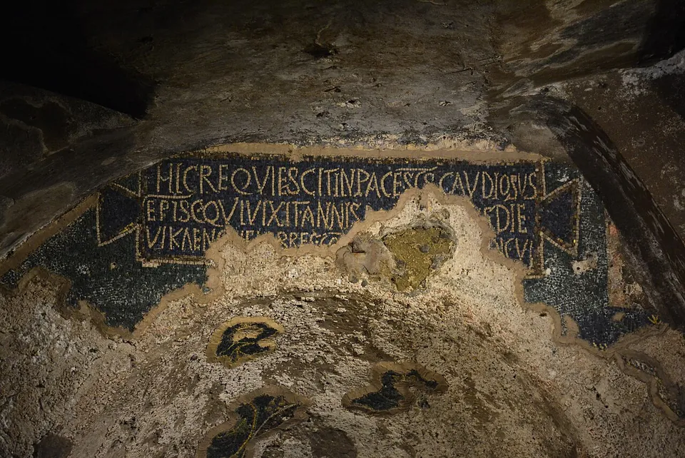 The top of an arched burial niche, where a band of mosaic in gold letters on dark blue begins HIC REQUIESCIT IN PACE and names GAUDIOSUS, broken by a loss in the middle; below it most of the mosaic has fallen away, leaving a few dark fragments on bare plaster.