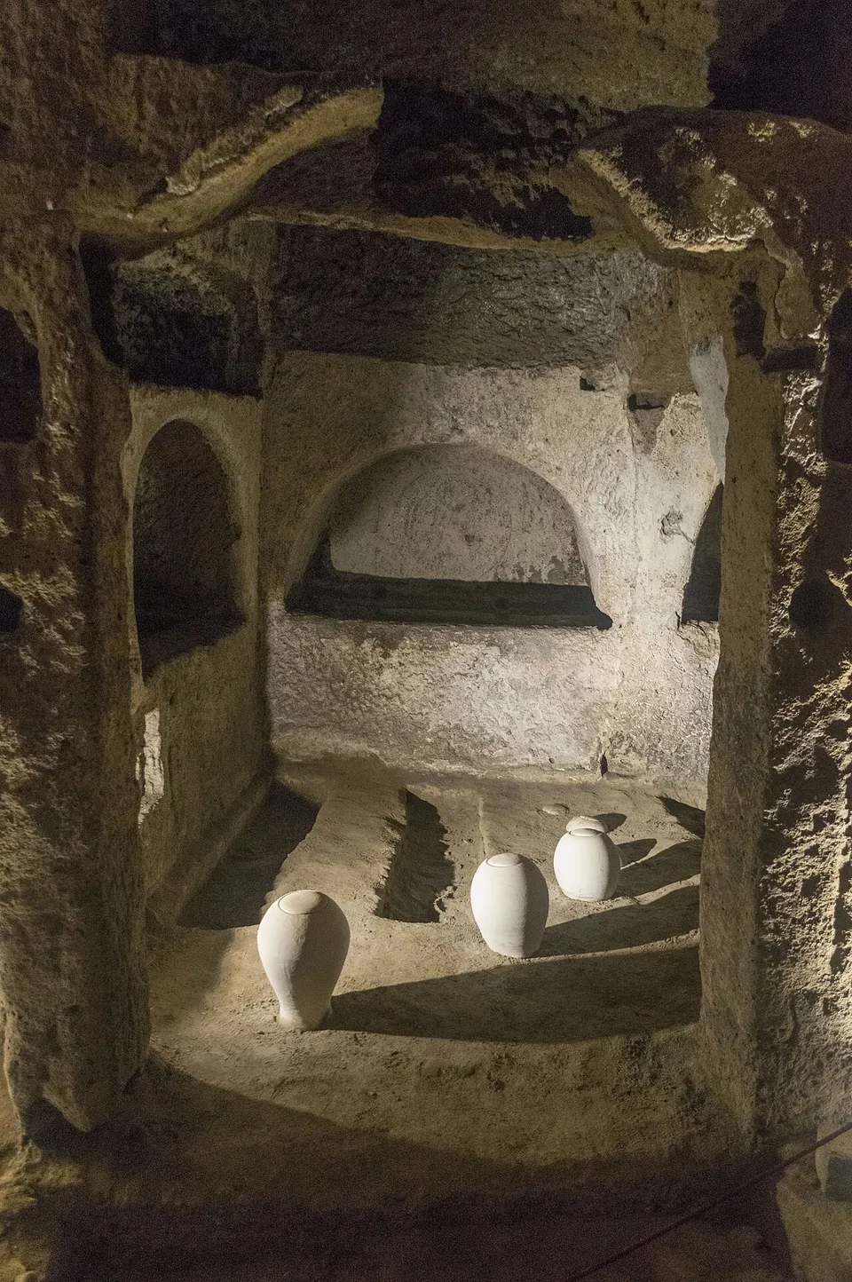 A broad gallery with a high, rounded ceiling, its rock walls cut into tiers of long horizontal niches, with tall arched openings into side chambers and lamps along the floor.