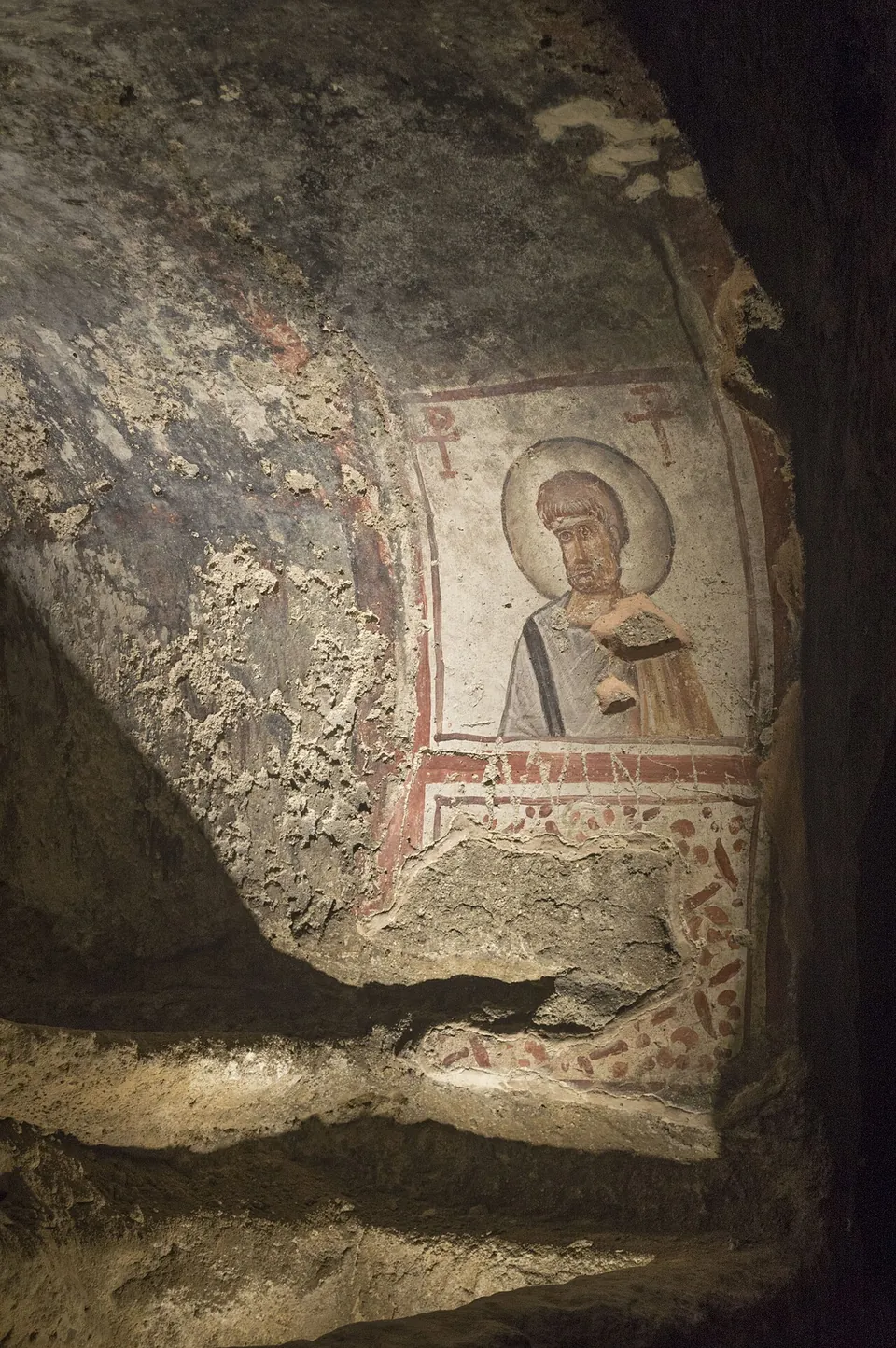 On a curved, flaking rock wall, a painted bust of a bearded man with a halo, in a white and grey robe, framed by red lines, with a cross symbol in each upper corner and a worn painted inscription below.