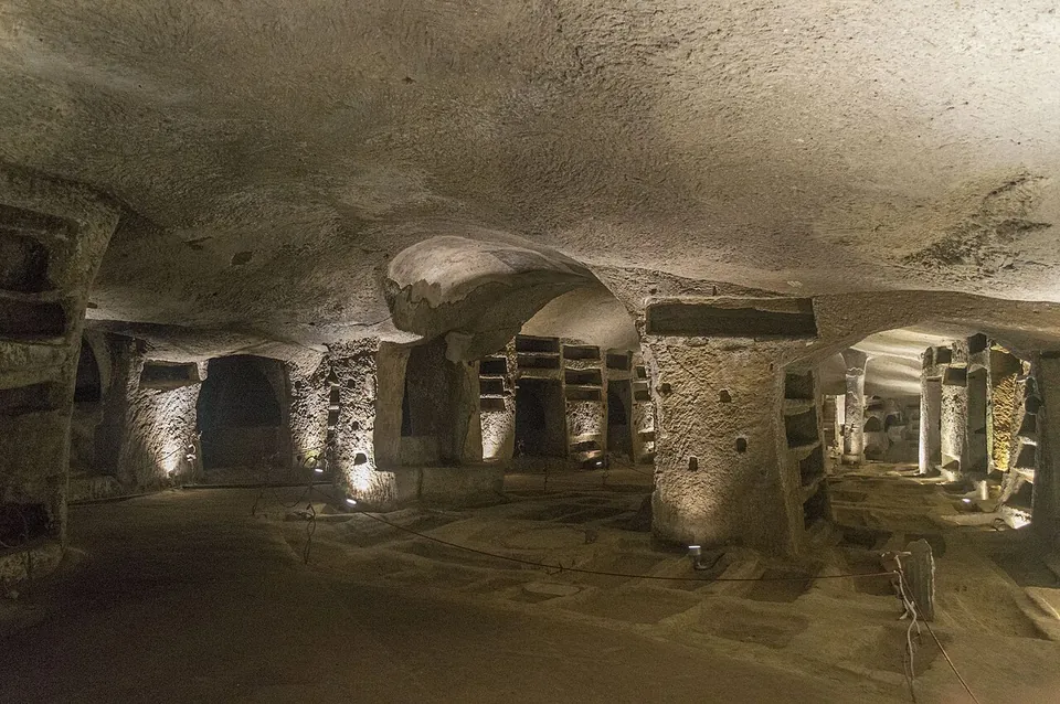 A wide, low hall cut into pale tuff, its flat ceiling held up by thick piers of living rock that are lined with rows of empty burial niches, lit by small lamps set on the floor.