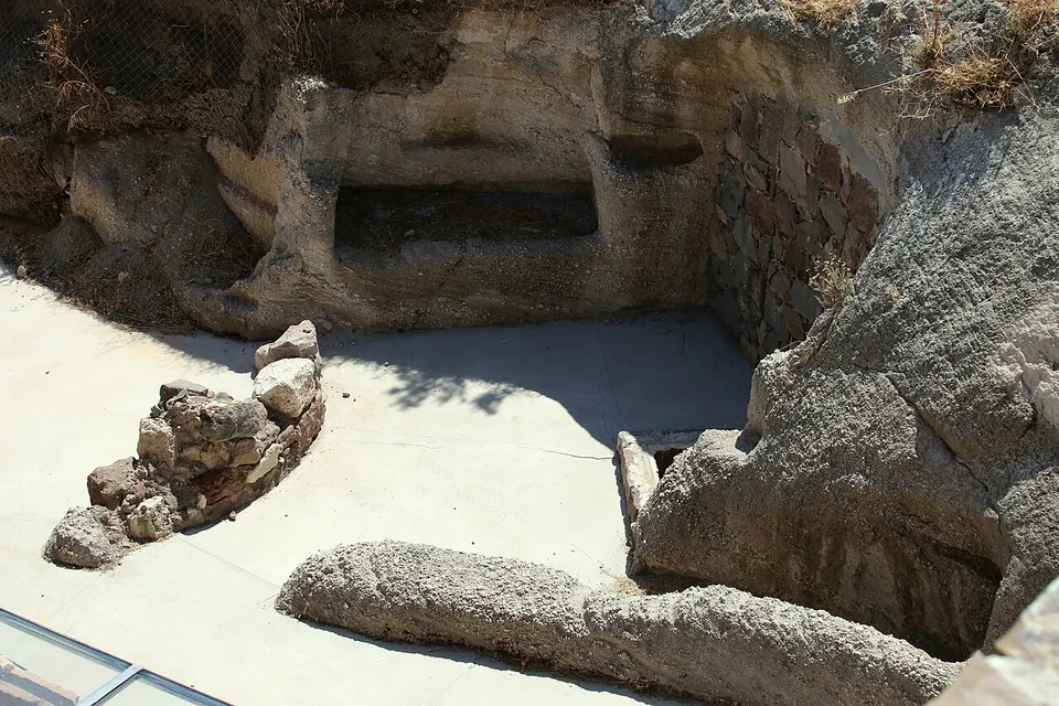 An open pit in grey volcanic rock with a long rectangular recess cut into its back wall, a short stretch of rubble wall and a rock-cut trough in the foreground.