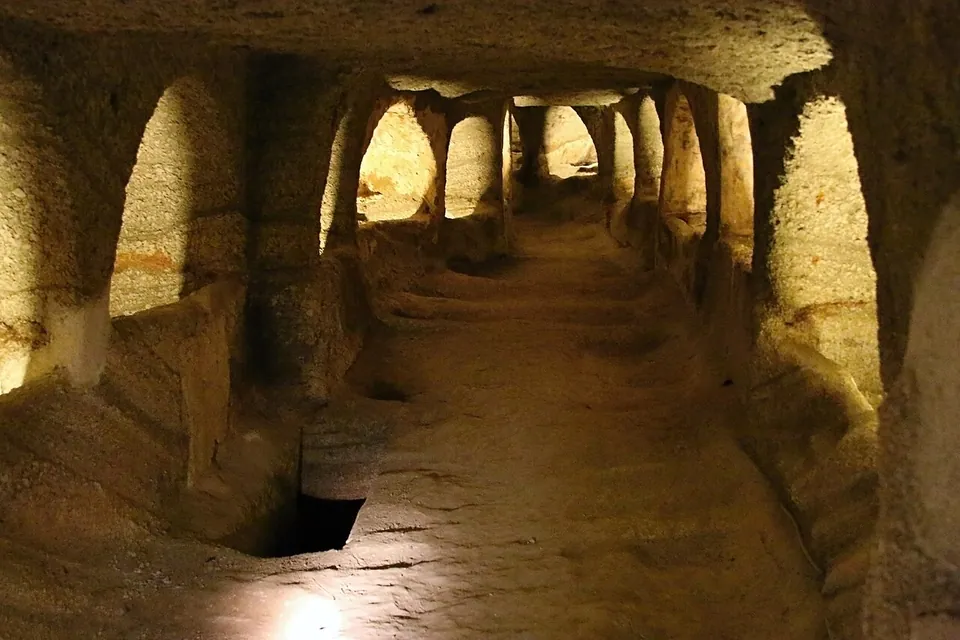A long underground corridor cut in pale volcanic rock, lit from below, with arched burial recesses opening on both sides and a pit grave cut into the floor.