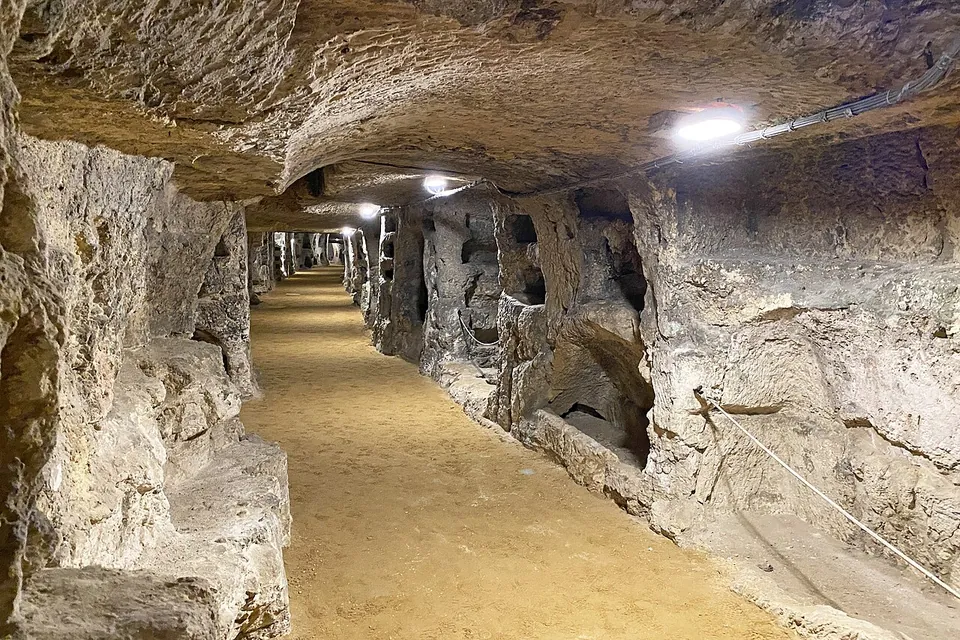A long, straight gallery cut through pale rock, with a sandy floor and electric lamps along the ceiling, its walls opened up by arched tomb recesses and niches on both sides.