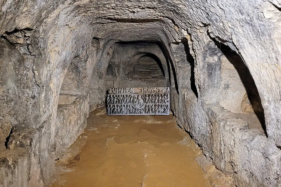 A rock-cut gallery with a sandy floor ending in a dark carved relief panel behind a low metal grille, with arched tomb recesses on either side.