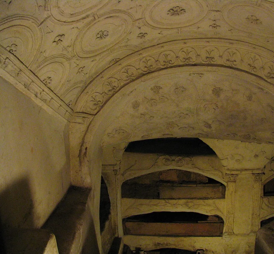 A low vault covered in pale stucco, moulded into roundels and bands of small leaf-like motifs, curving over a wall of stacked arched burial niches.