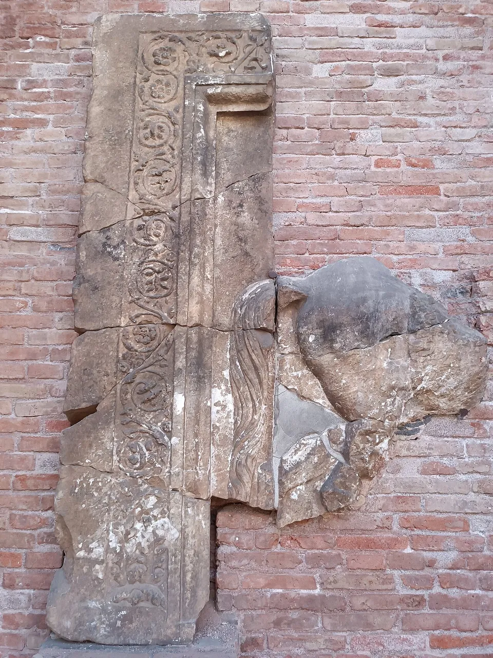 A weathered stone fragment carved with a band of spiral ornament, set upright against a wall of red brick next to a broken lump of stone.