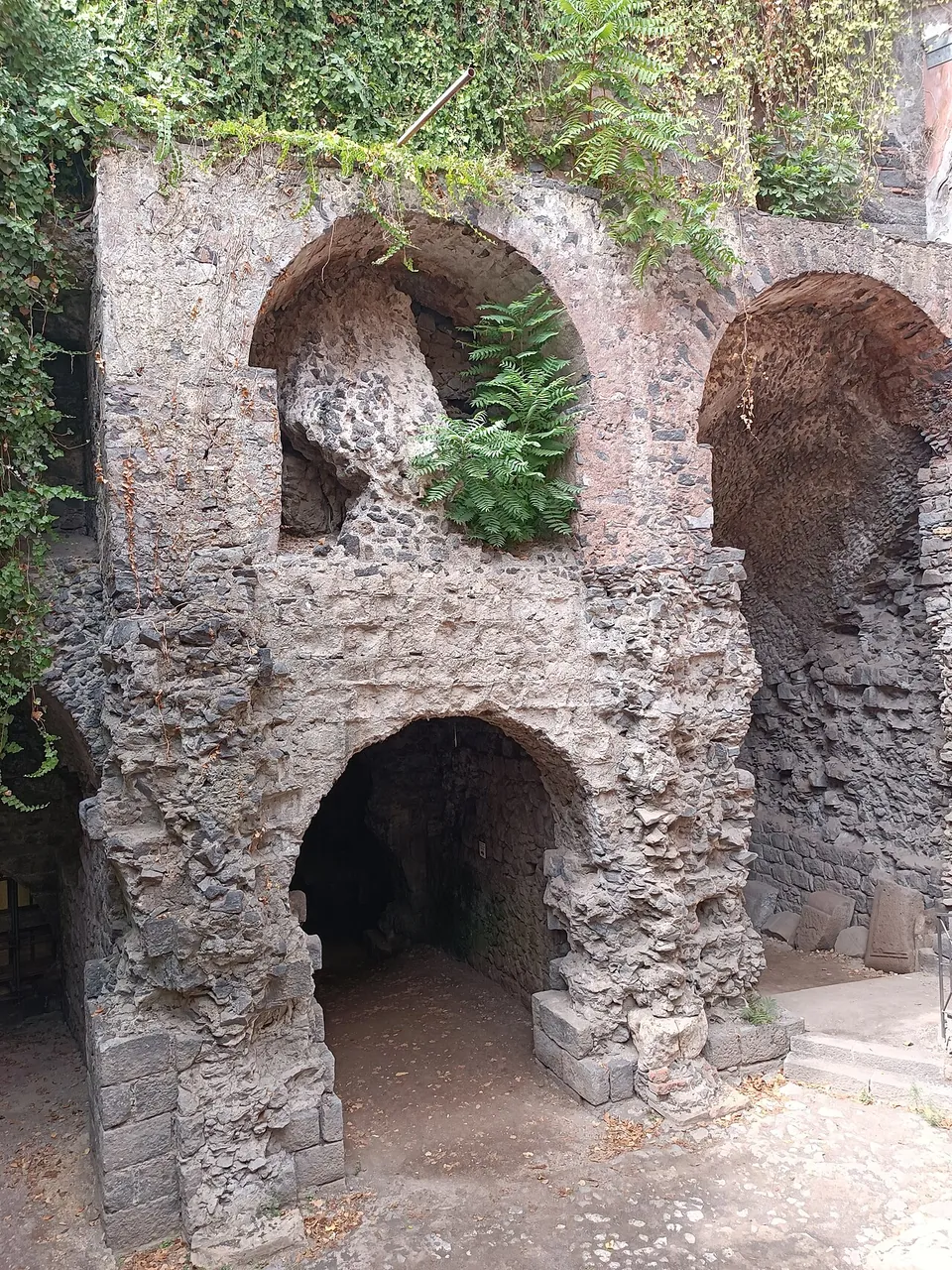 A ruined wall of rough grey and dark stone pierced by two levels of arched openings, with plants growing from the top.