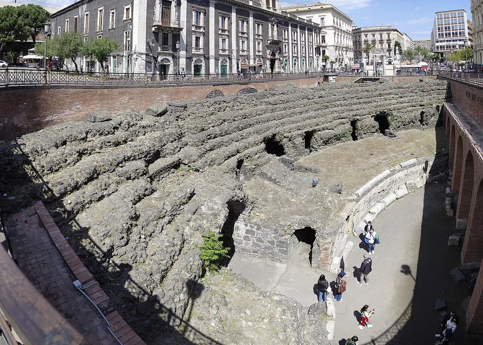 Looking down from street level into a sunken excavation where curved rows of black lava seating run around part of a sandy arena, with visitors walking on the sandy floor below and city buildings above.