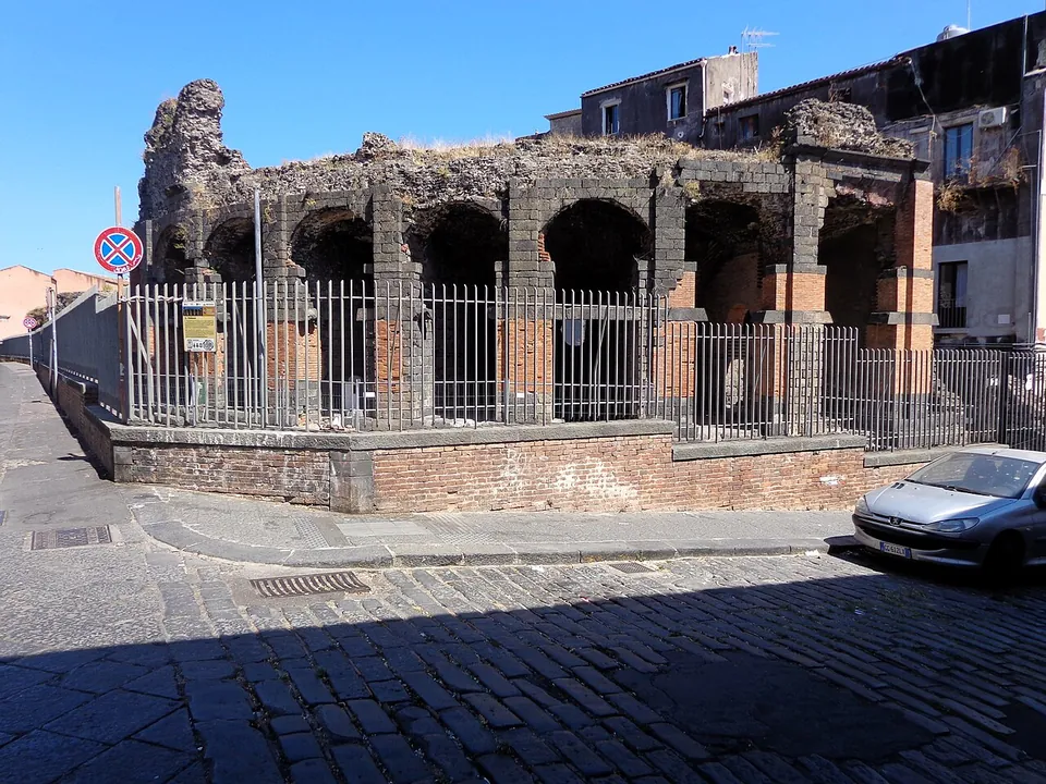 A cobbled street corner in sunshine with a railing on a low brick base, behind which a ruined arcade of dark stone with brick piers stands below apartment buildings; a parked car at the right.