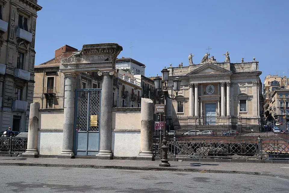 An iron gate between two marble columns carrying a carved architrave, with further column shafts at either end and low walls between them, standing at the edge of a square in front of a church façade with statues on its roofline.
