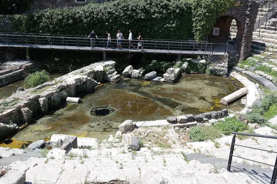 A shallow pool of greenish water lying over white stone paving and a fallen column, with visitors walking along a raised metal walkway above it and ivy on the wall behind.