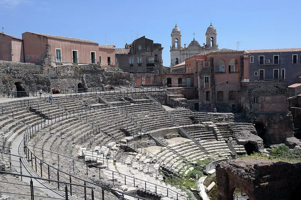 Curving tiers of dark stone seating with railed walkways across them, rising towards a row of old town houses and the twin bell towers of a church against a blue sky.