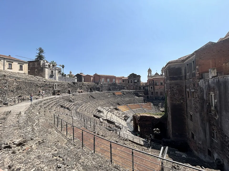 A wide view across the dark stone seating of the theatre, curving away to the right behind a railing, with the walls of houses built into its edge and a church tower and trees on the skyline.