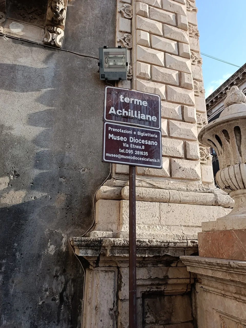 A brown street sign on a pole against a pale baroque stone façade, reading "terme Achilliane", with "Prenotazioni e Biglietteria Museo Diocesano, Via Etnea 8" and a telephone number below.