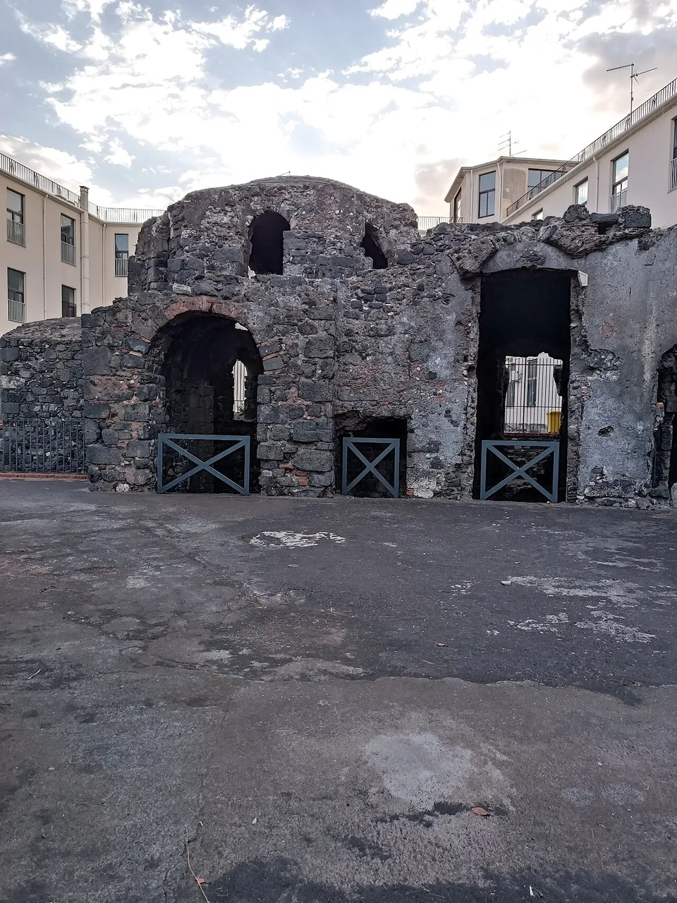 A paved open area of grey stone in front of the ruin, whose dark rubble walls rise to a low dome with arched openings, three of them at ground level closed by metal gates.