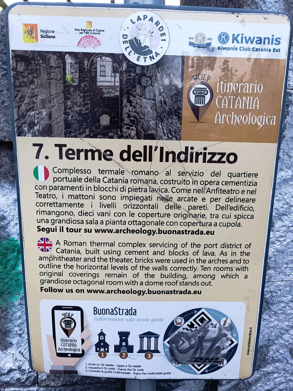 A bilingual information board headed "7. Terme dell'Indirizzo", in Italian and English, describing a Roman bath complex that served the port district, built of concrete faced with lava blocks, with ten rooms still roofed, among them a large octagonal domed hall; logos of the Sicilian Region and Kiwanis Club Catania Est at the top.