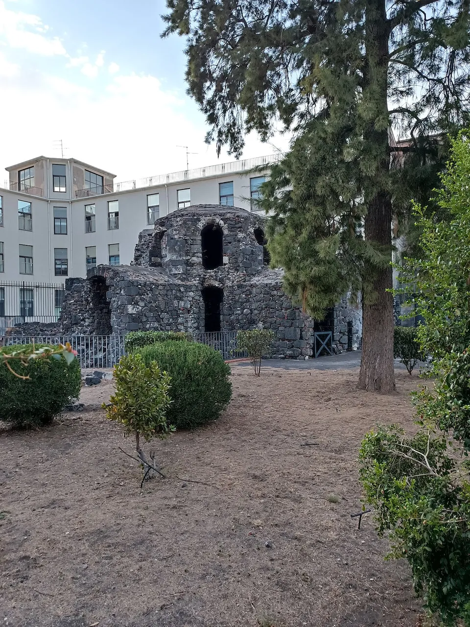 A dusty garden with clipped shrubs and a tall pine, and beyond a low railing a squat ruin of dark stone topped by a rounded dome with arched windows, in front of a white apartment block.