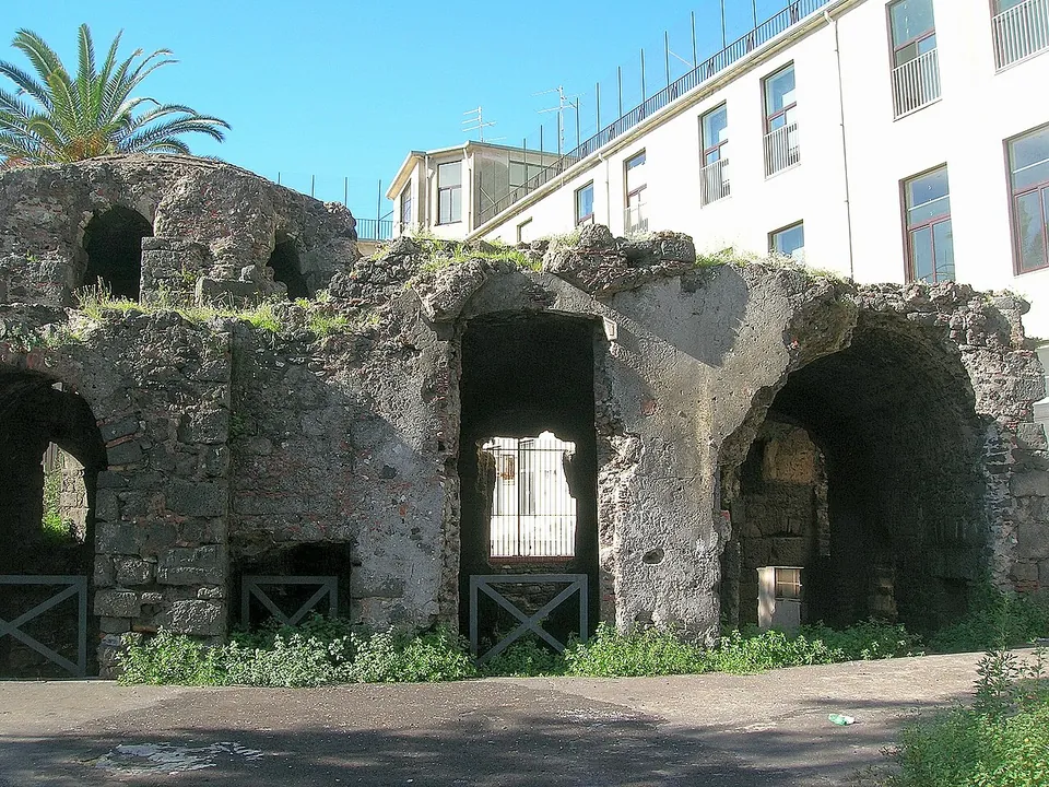 A long, low ruin of dark lava rubble with large arched openings fitted with low metal gates, grass growing on its broken vaults, a palm tree at the left and a white modern block rising behind.