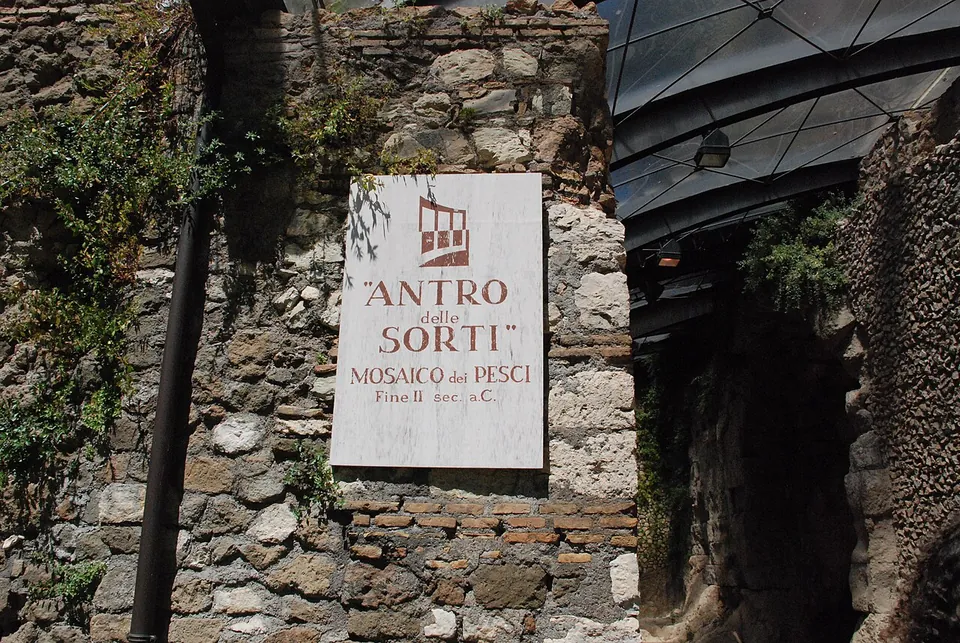 A white sign on a wall of rough stone and brick reads "Antro delle Sorti, Mosaico dei Pesci, Fine II sec. a.C.", beside a narrow passage under a glass roof.