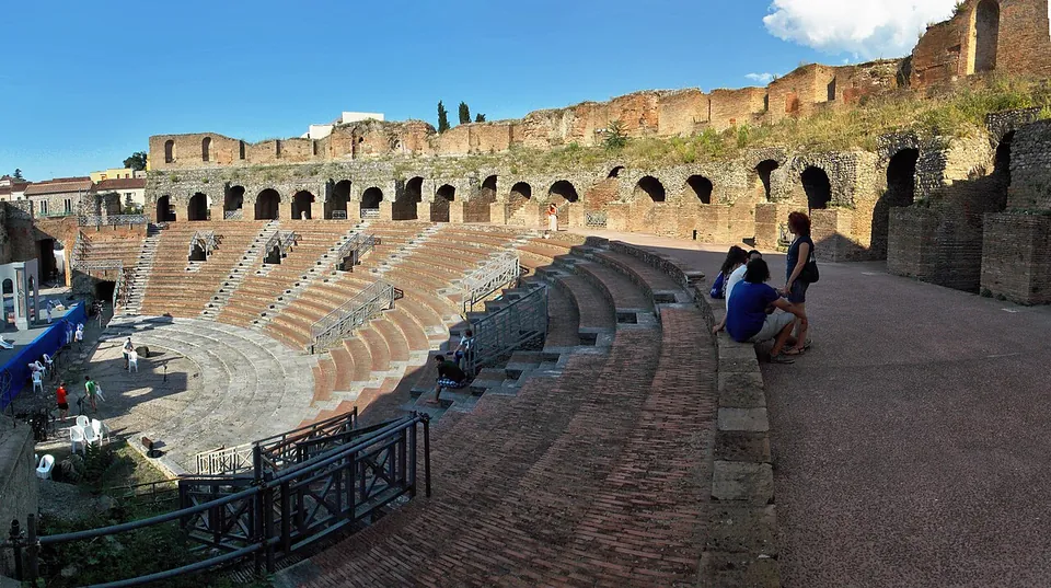 A semicircle of stone seating rising from a paved orchestra, with a ring of brick and stone arches standing along the top and modern railings and stage equipment at the edge; two visitors sit on the upper steps.