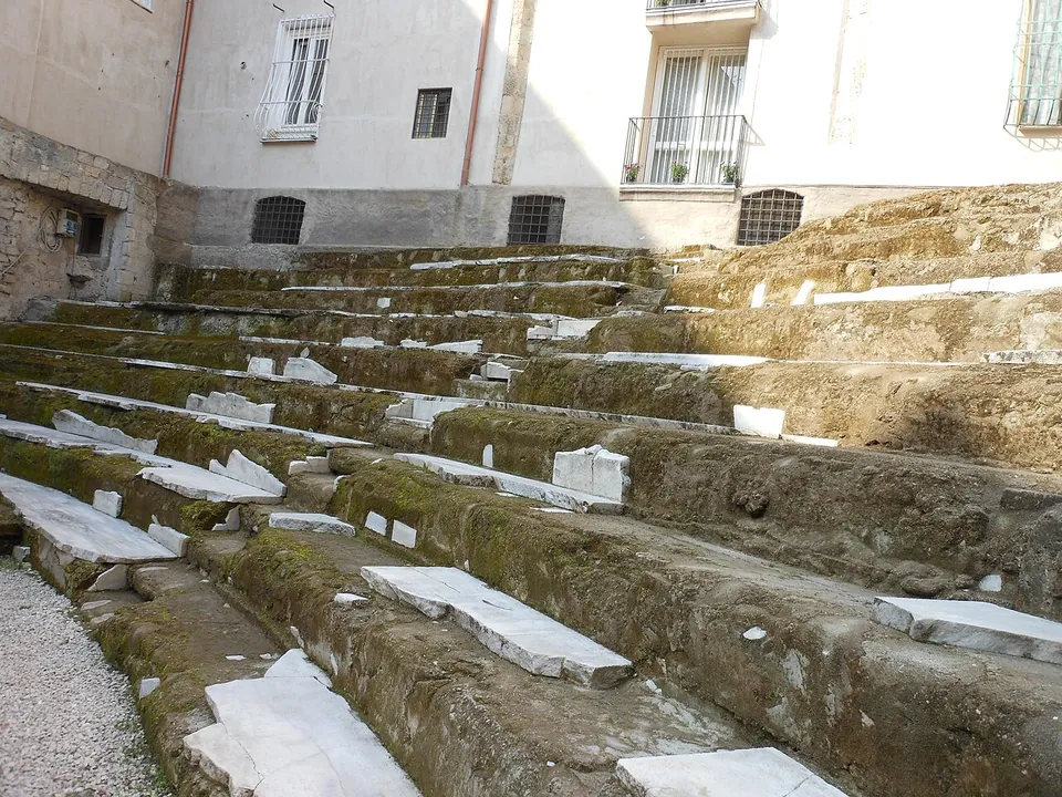 Curved rows of stone seating, some still faced with white marble slabs, in a courtyard overlooked by the windows and balconies of a later apartment building.