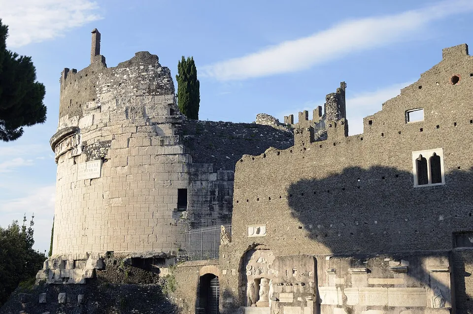 A massive drum of squared pale stone blocks with a ruined, uneven top stands beside a tall wall of darker rubble masonry with battlements and a pair of arched windows, under a blue sky.