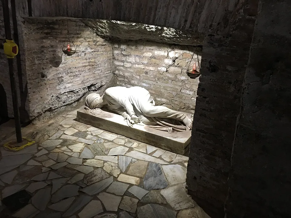 In a small underground chamber of old brick, a white marble statue of a young woman lies on her side on a low slab, her face turned to the floor, above a floor of irregular marble pieces, with hanging lamps on the wall.