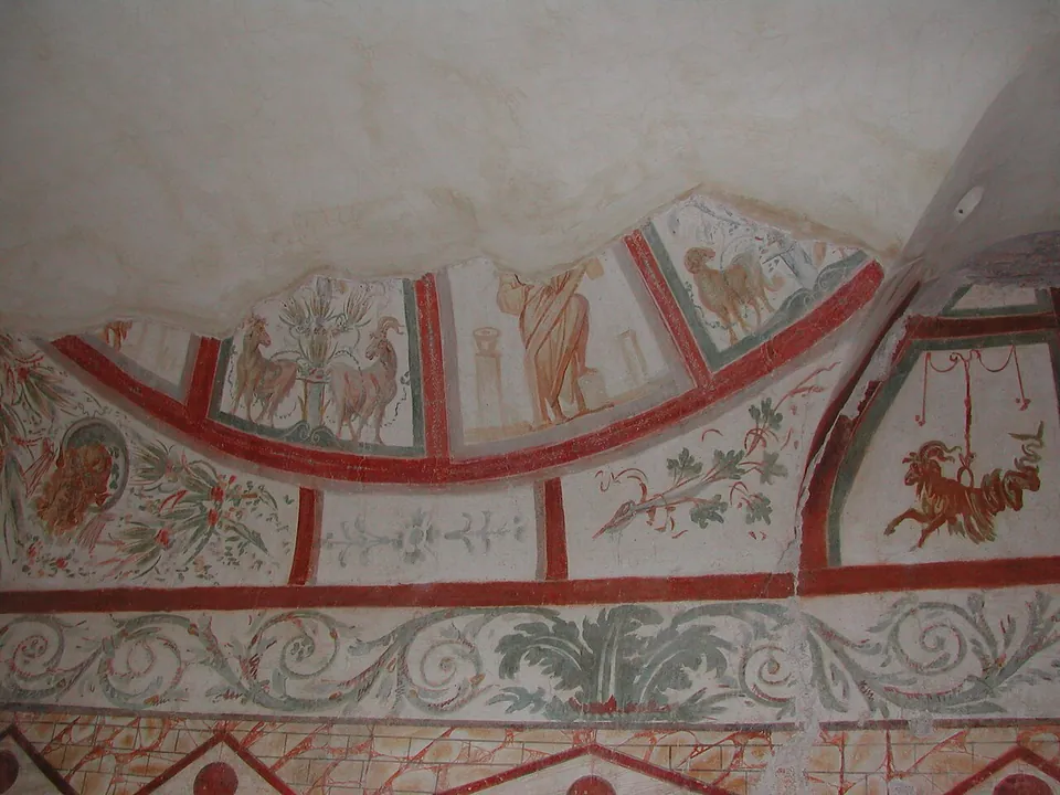 A painted vault divided by red bands into panels holding pairs of goats or rams beside a plant and a standing draped figure, with a band of green scrolling foliage below.