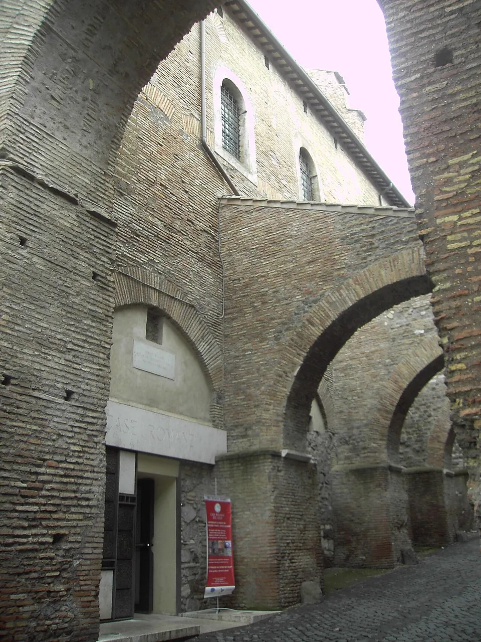 A doorway with a red banner beside it at the foot of a high brick wall, with brick arches springing across a cobbled lane from the wall.