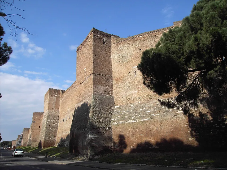 A tall brick wall with square towers at intervals running away beside a road under a blue sky, an umbrella pine in front of the nearest tower and a car further along the road.