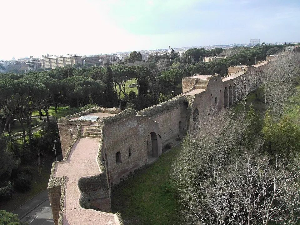 Looking down from a gate tower onto the top of the wall, a paved walkway between parapets running away past square towers through a belt of pine trees, with apartment blocks on the skyline.