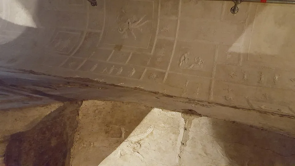 The barrel vault of the central aisle seen from below, its white stucco divided into rectangular and square panels with small figures in low relief, a scaffolding pipe running along one edge and the top of a pillar arch in the lower corner.