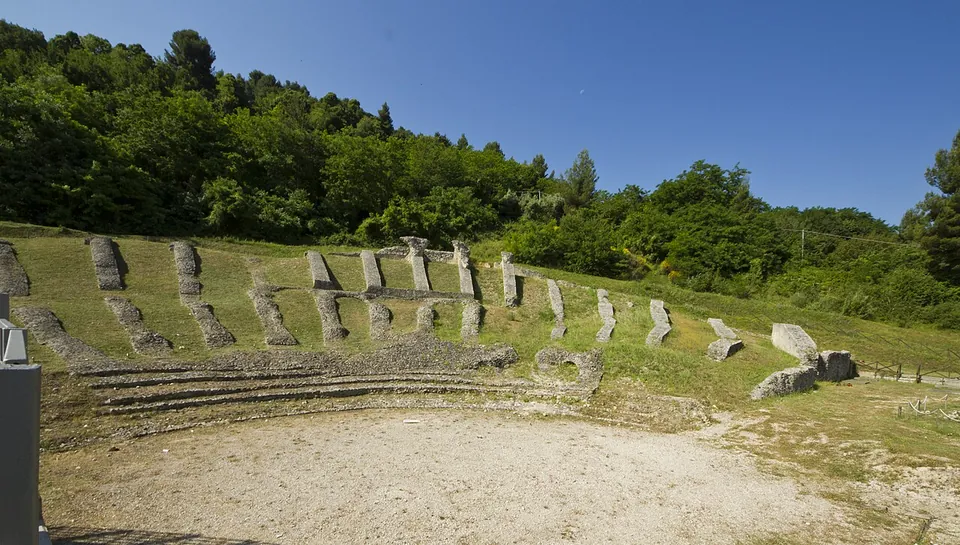 A semicircle of short stone wall stubs set in mown grass climbs a slope towards a wooded hill, around a level floor of packed earth, under a clear blue sky.