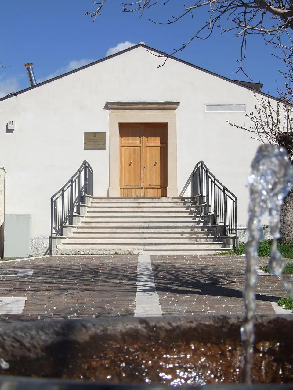 A plain white building with a tall wooden door at the top of a flight of stone steps with metal handrails, seen across a paved square past a fountain spout.