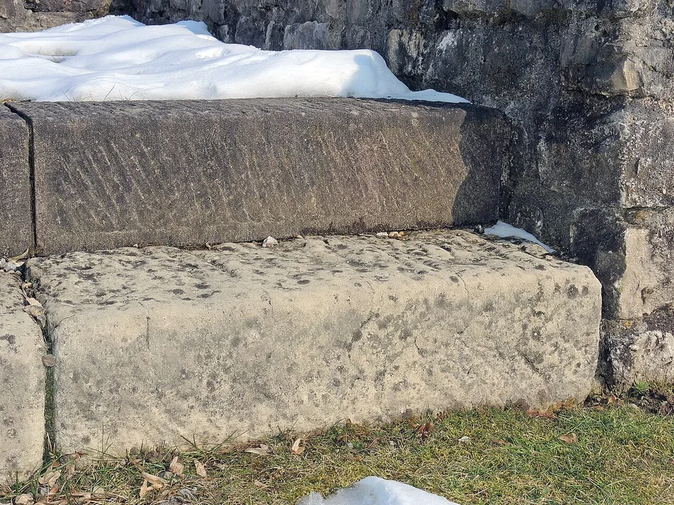 Two large dressed stone blocks, one laid on the other, the upper one marked with diagonal tool lines, against a rubble wall with snow lying on it.