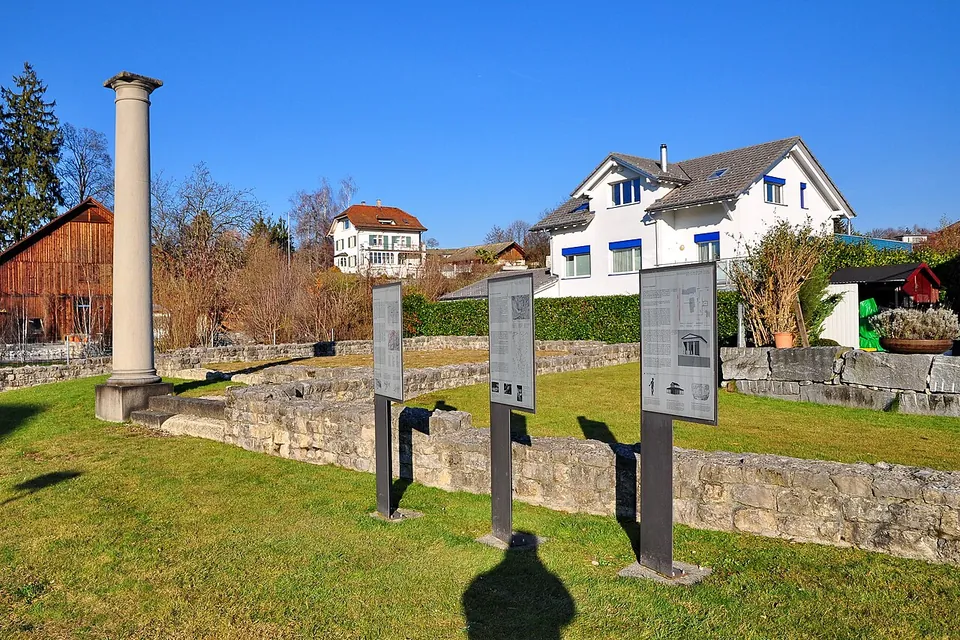 Low stone foundation walls laid out on a lawn, with a single column standing at one corner and three information panels on posts, among houses.