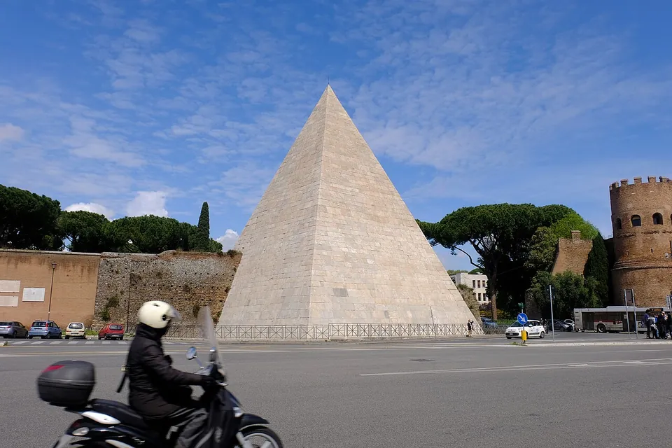 A steep white marble pyramid seen across a busy square, with a stretch of old brick wall and umbrella pines to its left and a round crenellated brick tower to its right; a motor scooter crosses the foreground.