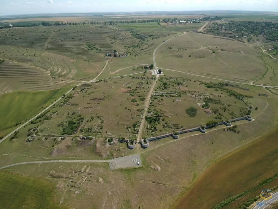 An aerial view over open grassland of a walled Roman town. A wall with rounded towers runs across the foreground, a straight road crosses the enclosure, and low excavated foundations are scattered inside, with farmland and a village beyond.