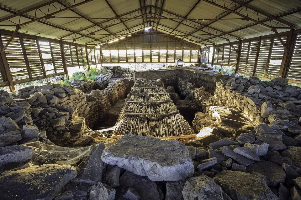Excavated stone walls and rubble under a large open-sided shelter of timber and corrugated roofing, with a long trench lined with reed matting down the middle.