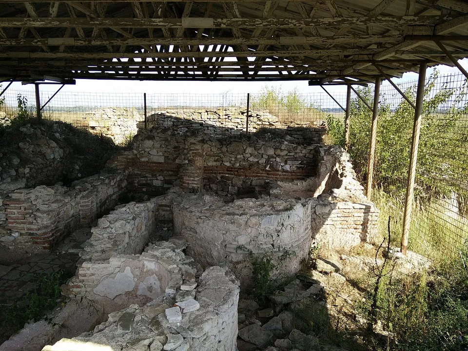 Walls of stone and brick with rounded basins and low rooms, under a metal roof on posts, with a fence and scrub beyond.