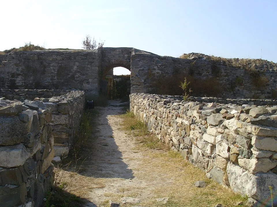 A narrow passage of beaten earth between walls of rough stone, leading to an arched opening in a higher wall, under a clear sky.
