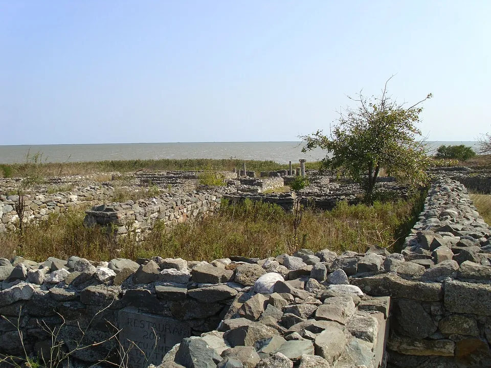 Rough stone walls of small rooms and courtyards overgrown with dry grass, with a bush and the lake behind them.