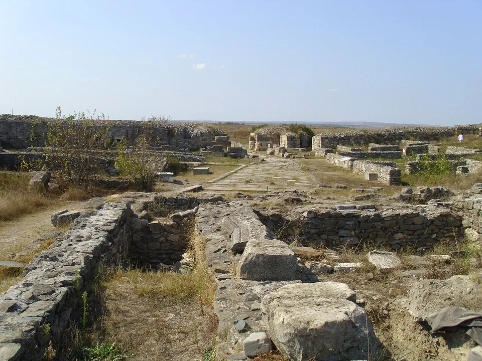 Low stone walls and fallen blocks of an excavated building in dry grass, with a paved street and more ruins stretching to the city wall on the horizon.