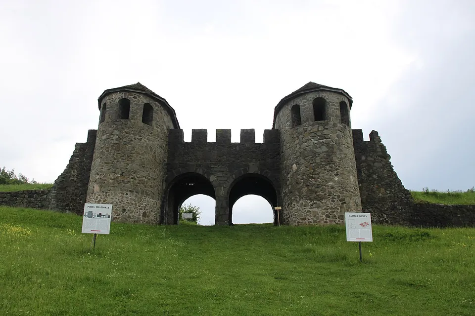 A rebuilt Roman gate seen from the front across a lawn, two round-fronted stone towers with small windows flanking a crenellated wall pierced by two arched passages, with information boards in front.