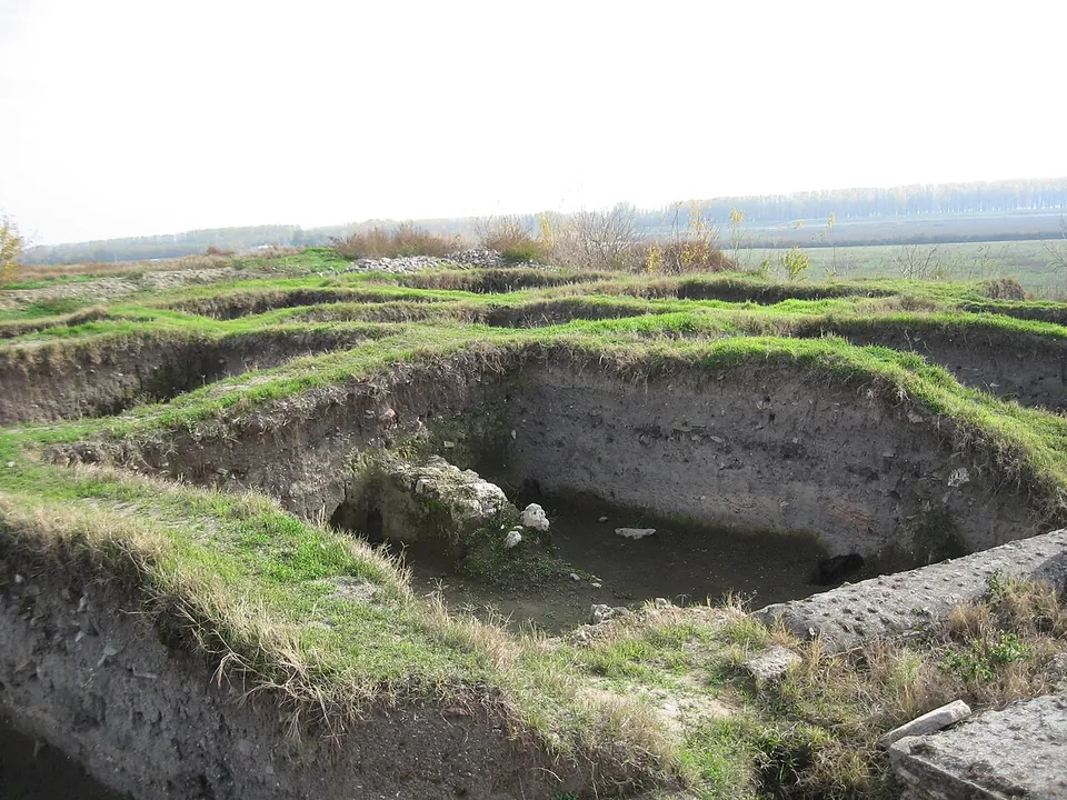 Grass-grown excavation trenches and low stone walls on a terrace, with the flat plain of the Danube stretching to a line of trees on the horizon.