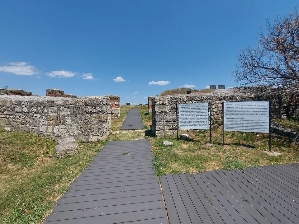 A plank walkway leading to a gap between two stretches of stone wall, with two information boards standing in front of the wall under a blue sky.