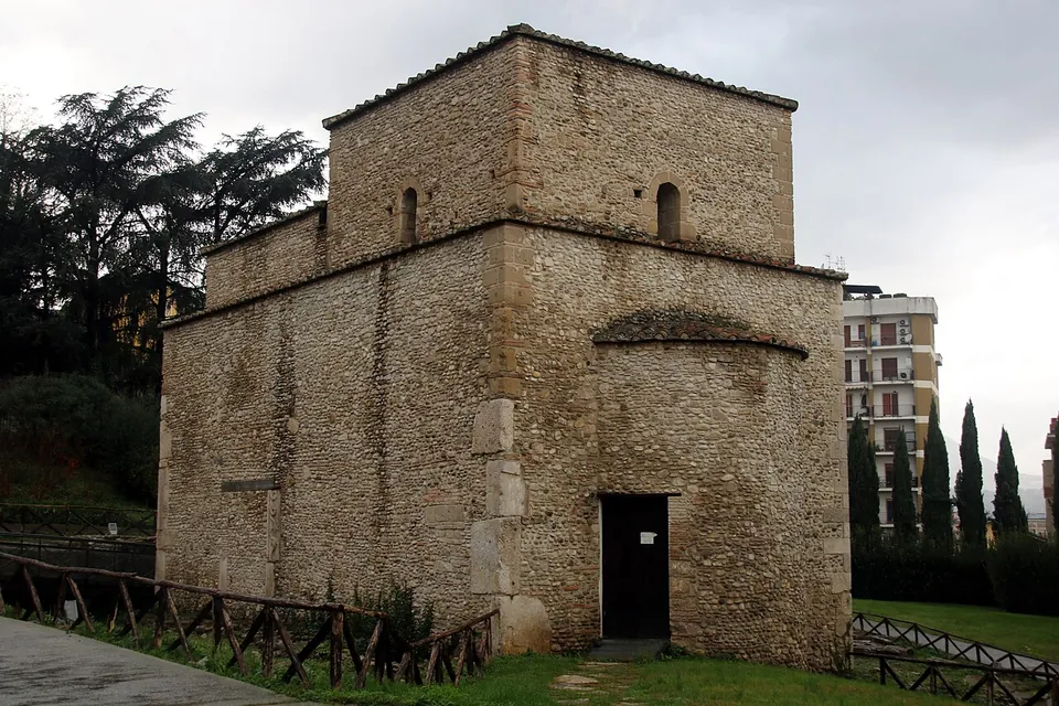 A small, plain building of rough stone with a square upper storey and a single doorway, standing on a lawn beside a low wooden fence, with trees and blocks of flats behind.